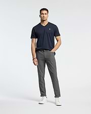 A man stands against a white background wearing gray pants and the Psycho Bunny MENS CLASSIC V NECK TEE - B6U100ARPC in dark navy. Made of Pima cotton, it pairs well with his short curly hair and white sneakers. His right hand is in his pocket, with his left arm by his side.