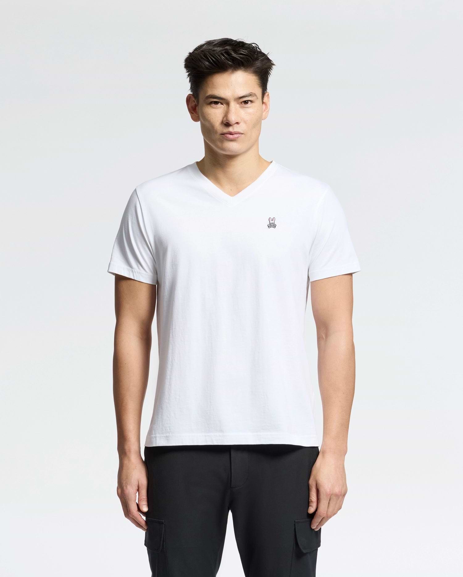A man with short dark hair is wearing a white MENS CLASSIC V NECK TEE - B6U100ARPC by Psycho Bunny, made in Peru from 100% Pima cotton, featuring a small emblem on the chest. He pairs it with black pants and stands against a light gray background.