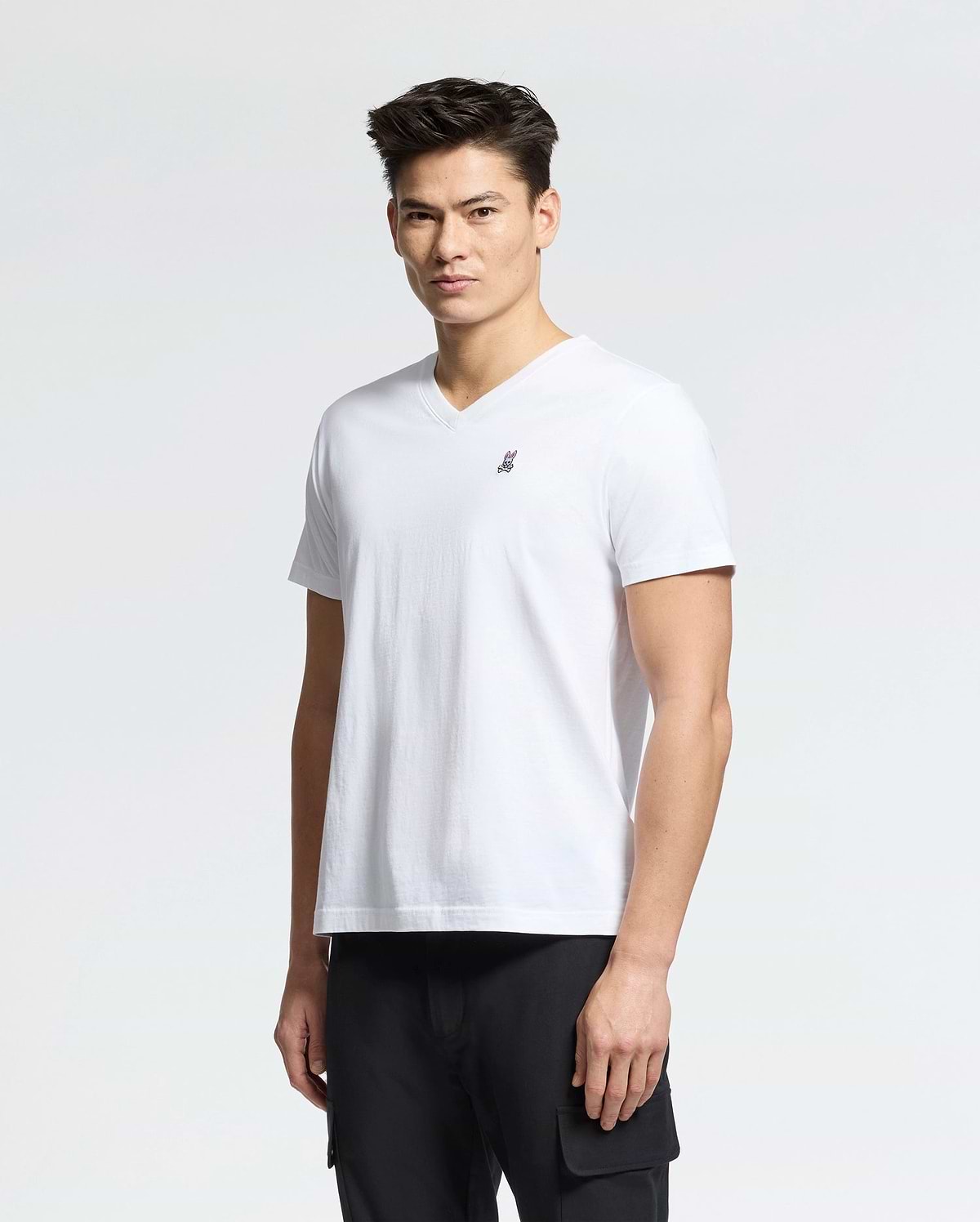 A person with short hair is wearing a Psycho Bunny MENS CLASSIC V NECK TEE - B6U100ARPC made from 100% Pima cotton, produced in Peru, featuring a small embroidered rabbit logo on the chest. They're pairing it with black pants and standing against a plain white background.