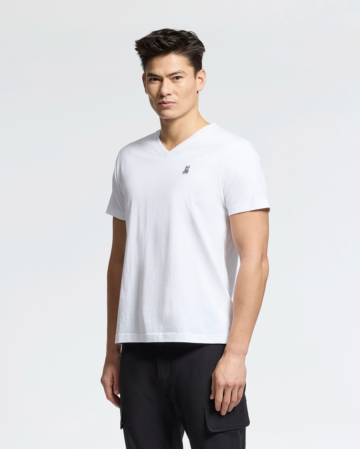 A person with short hair is wearing a Psycho Bunny MENS CLASSIC V NECK TEE - B6U100ARPC made from 100% Pima cotton, produced in Peru, featuring a small embroidered rabbit logo on the chest. They're pairing it with black pants and standing against a plain white background.