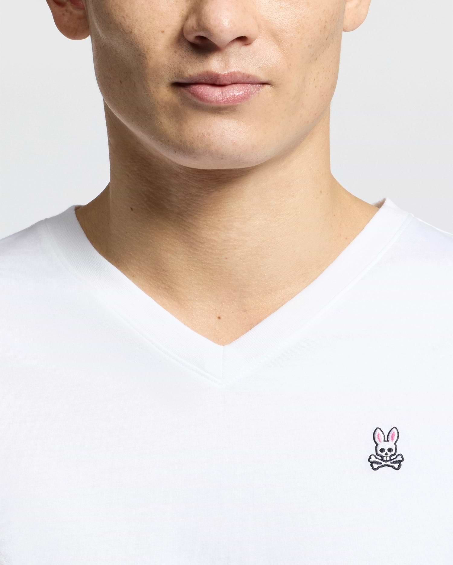 Close-up of someone wearing the Psycho Bunny MENS CLASSIC V NECK TEE - B6U100ARPC, made in Peru from 100% Pima cotton. The shirt has a white background with a small embroidered pink bunny above crossed bones on the chest.
