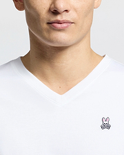 Close-up of someone wearing the Psycho Bunny MENS CLASSIC V NECK TEE - B6U100ARPC, made in Peru from 100% Pima cotton. The shirt has a white background with a small embroidered pink bunny above crossed bones on the chest.