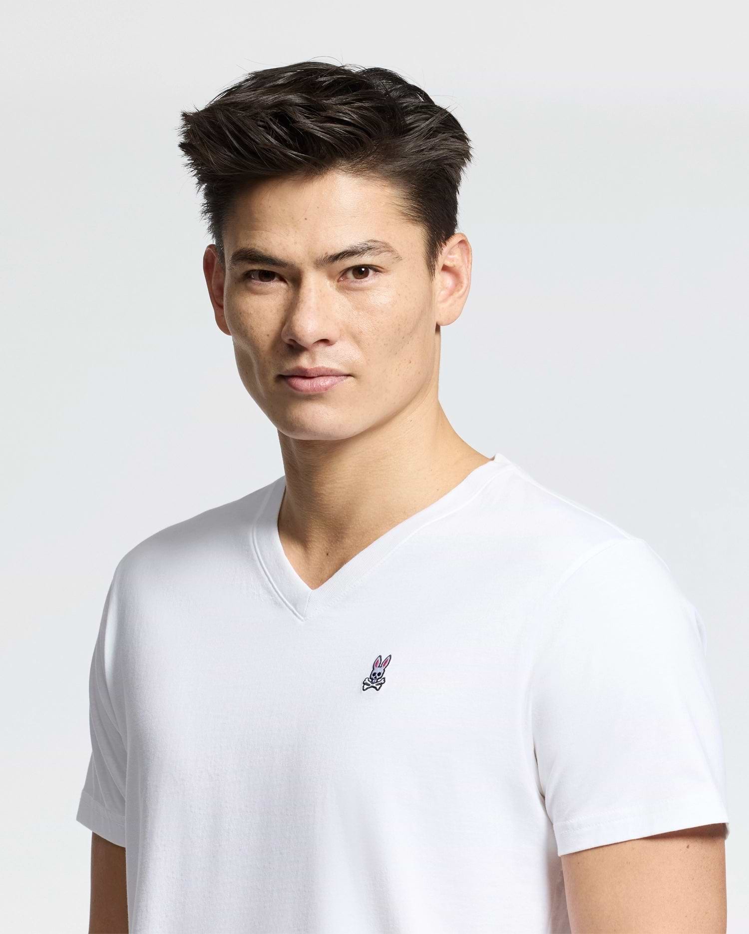 A person with short dark hair wears a white Psycho Bunny MENS CLASSIC V NECK TEE - B6U100ARPC, crafted in Peru from 100% Pima cotton, with a small chest logo. They face the camera against a plain gray background.
