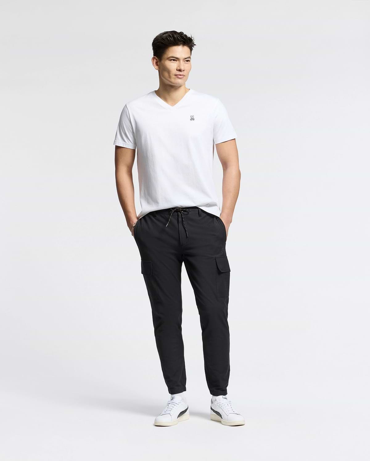 A man stands against a plain white background, wearing the MENS CLASSIC V NECK TEE - B6U100ARPC by Psycho Bunny, crafted in Peru. Paired with black cargo pants and white sneakers, his hands are in his pockets as he looks to the side, exuding relaxed confidence in 100% Pima cotton.