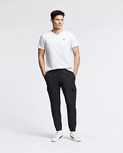 A man stands against a plain white background, wearing the MENS CLASSIC V NECK TEE - B6U100ARPC by Psycho Bunny, crafted in Peru. Paired with black cargo pants and white sneakers, his hands are in his pockets as he looks to the side, exuding relaxed confidence in 100% Pima cotton.