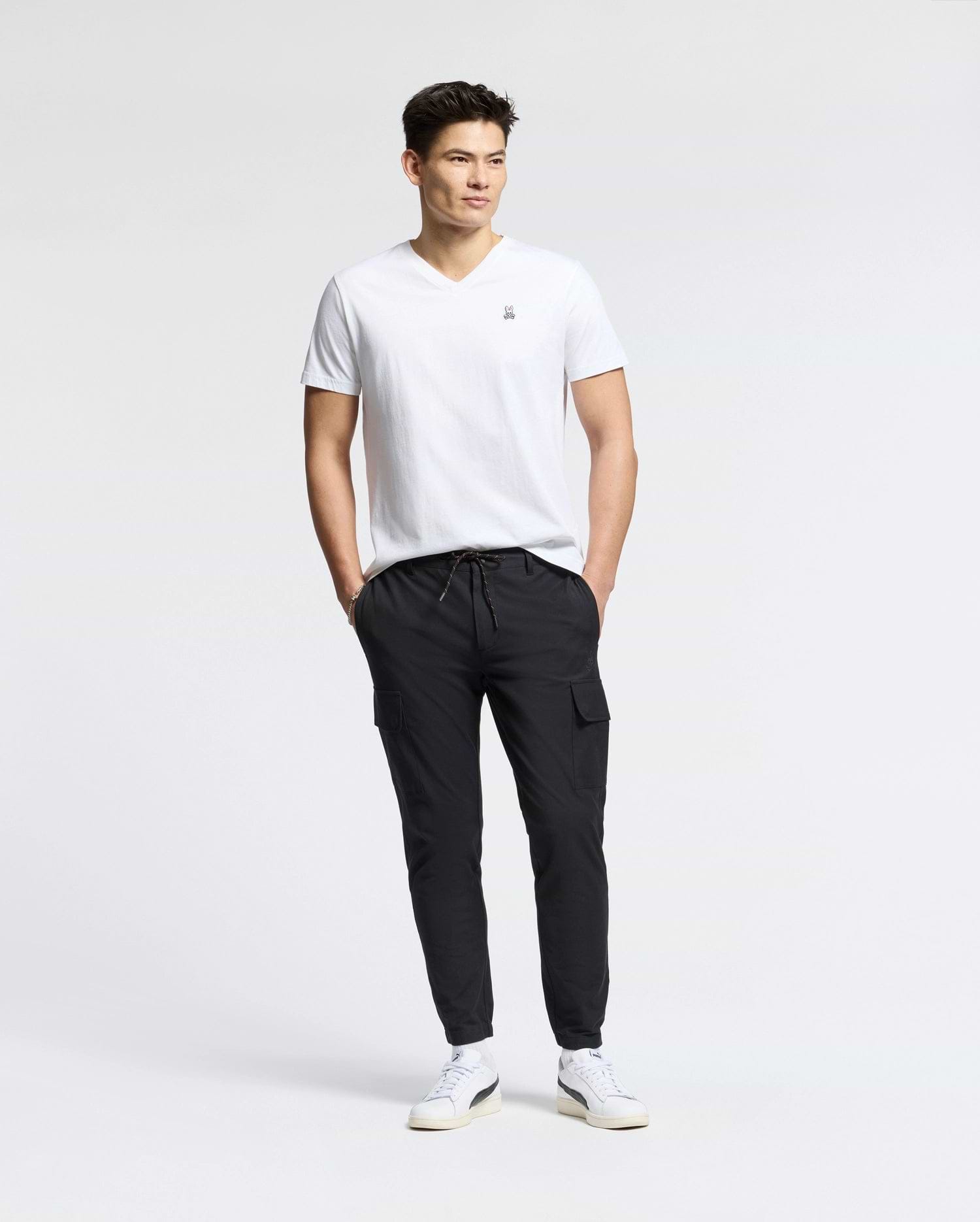 A man stands against a plain white background, wearing the MENS CLASSIC V NECK TEE - B6U100ARPC by Psycho Bunny, crafted in Peru. Paired with black cargo pants and white sneakers, his hands are in his pockets as he looks to the side, exuding relaxed confidence in 100% Pima cotton.