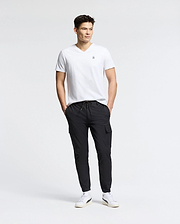 A man stands against a plain white background, wearing the MENS CLASSIC V NECK TEE - B6U100ARPC by Psycho Bunny, crafted in Peru. Paired with black cargo pants and white sneakers, his hands are in his pockets as he looks to the side, exuding relaxed confidence in 100% Pima cotton.