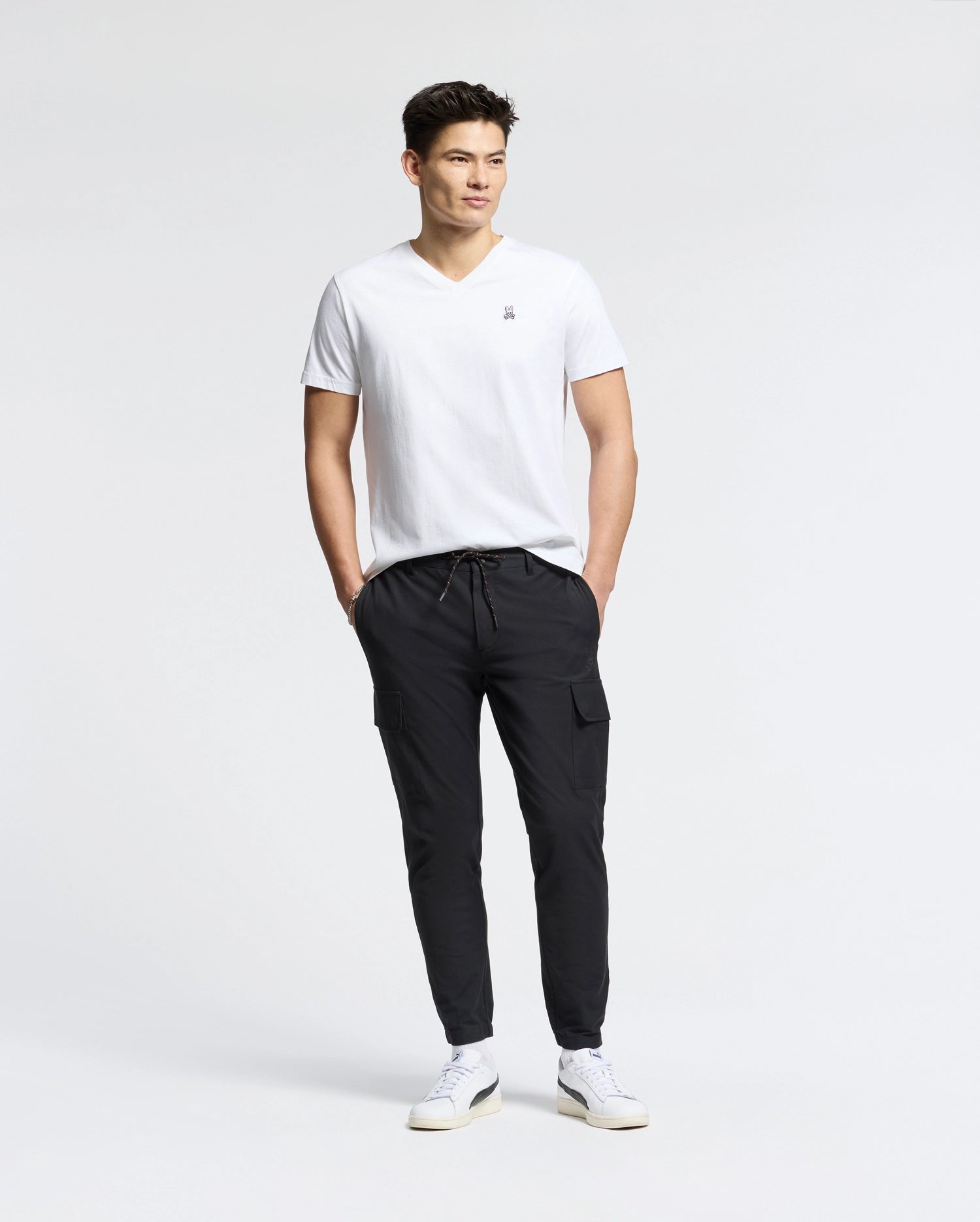 A man stands against a plain white background, wearing the MENS CLASSIC V NECK TEE - B6U100ARPC by Psycho Bunny, crafted in Peru. Paired with black cargo pants and white sneakers, his hands are in his pockets as he looks to the side, exuding relaxed confidence in 100% Pima cotton.