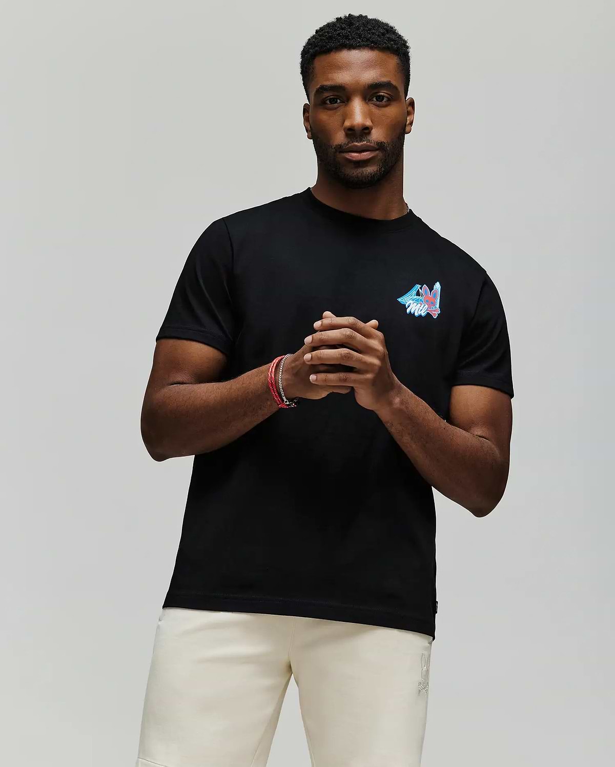 A man stands against a plain background wearing the Psycho Bunny MENS MONTREAL TEE (B6U381F200), a soft black Pima cotton t-shirt with a small colorful chest design, paired with light shorts. He clasps his hands and looks at the camera.