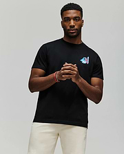 A man stands against a plain background wearing the Psycho Bunny MENS MONTREAL TEE (B6U381F200), a soft black Pima cotton t-shirt with a small colorful chest design, paired with light shorts. He clasps his hands and looks at the camera.