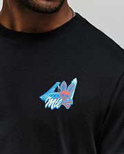A man wears the Psycho Bunny MENS MONTREAL TEE (B6U381F200), a black Pima cotton shirt with a small chest graphic featuring a pink Psycho Bunny skull and crossbones, blue geometric shapes, and “Mil” stylized text.