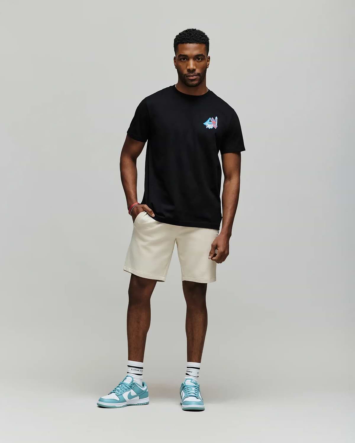 A man with short curly hair and a relaxed, confident posture wears the Psycho Bunny MENS MONTREAL TEE (B6U381F200), styled with beige shorts, white socks, and blue sneakers, embodying Montreal street style against a plain background.
