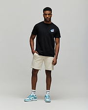 A man with short curly hair and a relaxed, confident posture wears the Psycho Bunny MENS MONTREAL TEE (B6U381F200), styled with beige shorts, white socks, and blue sneakers, embodying Montreal street style against a plain background.