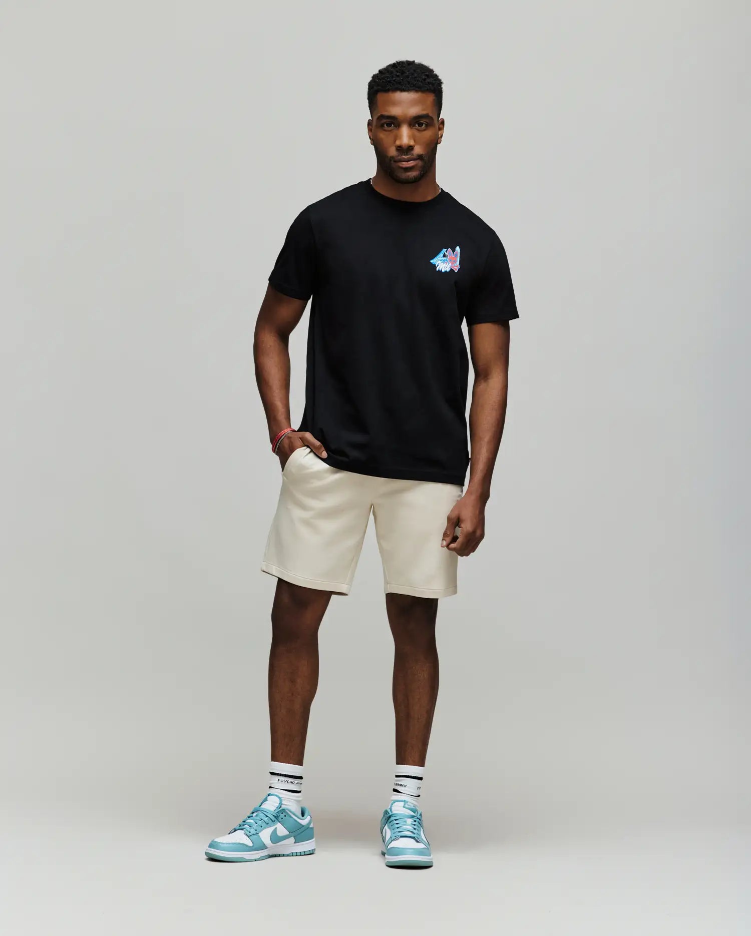 A man with short curly hair and a relaxed, confident posture wears the Psycho Bunny MENS MONTREAL TEE (B6U381F200), styled with beige shorts, white socks, and blue sneakers, embodying Montreal street style against a plain background.