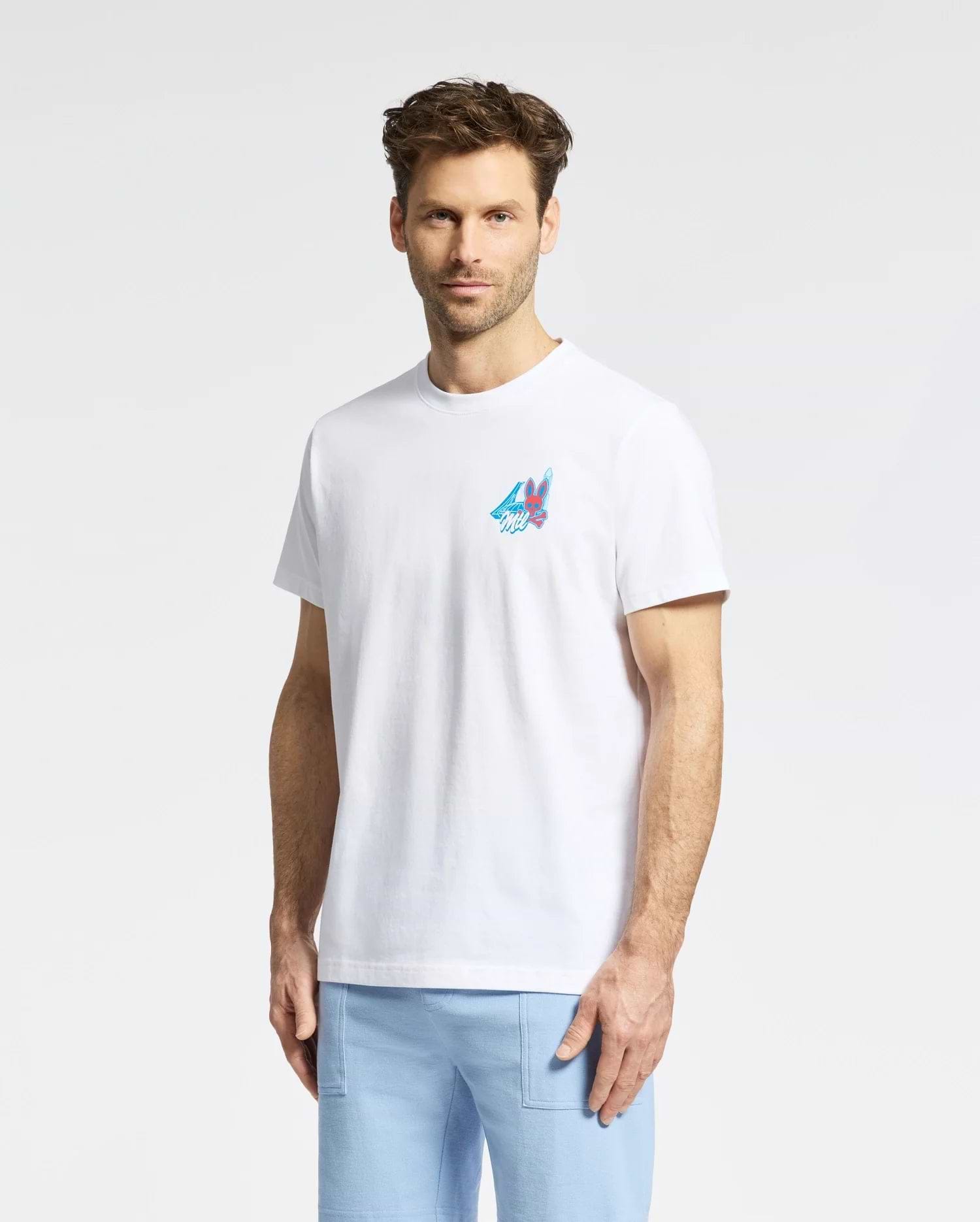 A man with short brown hair and a beard wears the Psycho Bunny MENS MONTREAL TEE - B6U381F200, featuring a small colorful mountain graphic on the left chest, and light blue pants, standing against a plain white background.