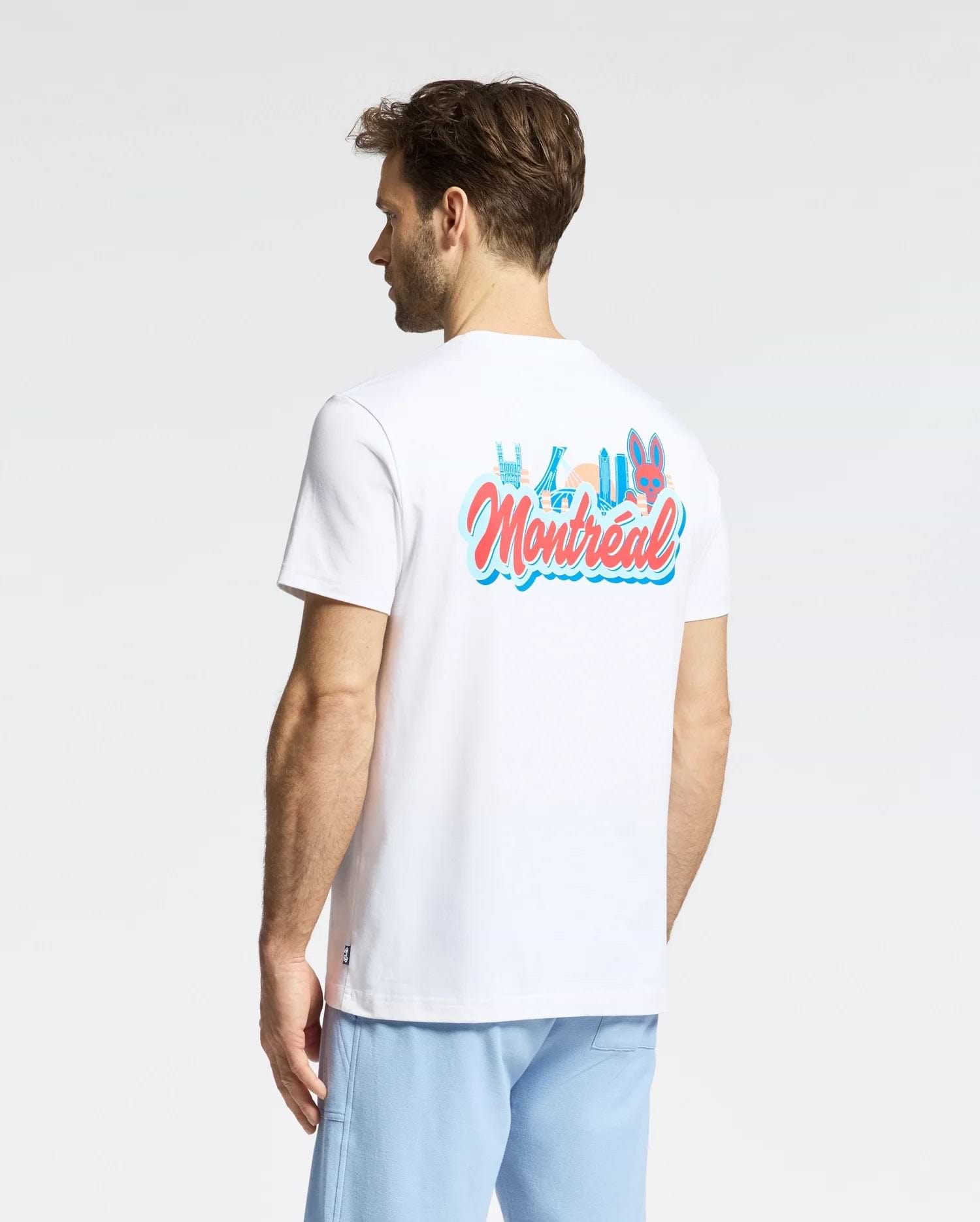 A man wearing the Psycho Bunny MENS MONTREAL TEE (B6U381F200), a white Pima cotton shirt with colorful "Montréal" text and a cartoon graphic on the back, stands facing away in light blue pants, displaying classic Psycho Bunny style.