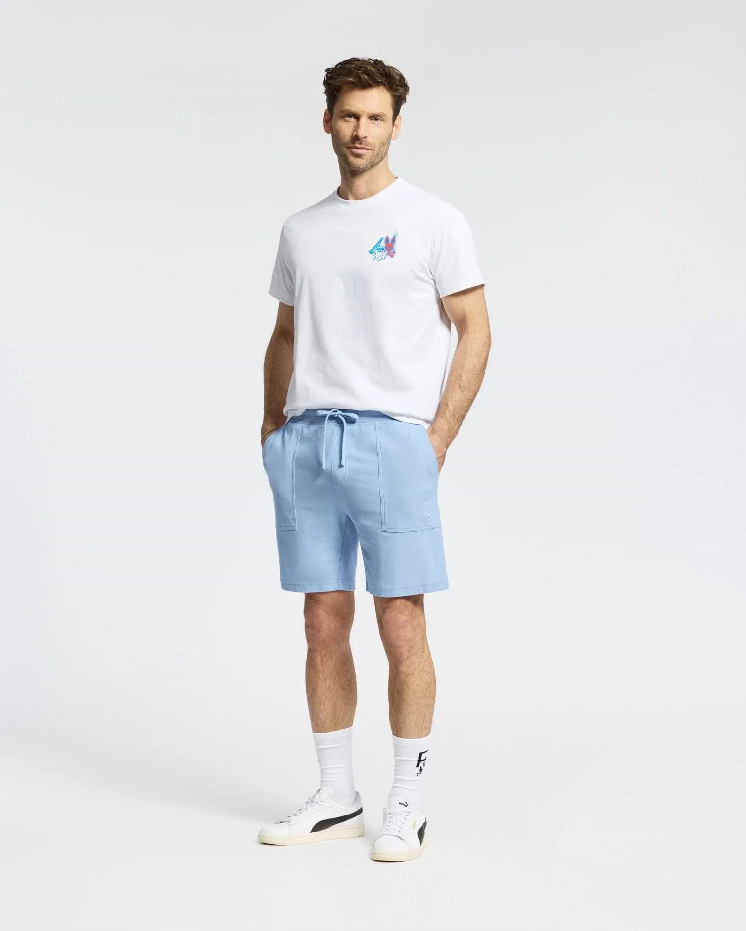 A man faces forward against a plain white background wearing a Psycho Bunny MENS MONTREAL TEE - B6U381F200 in white, paired with light blue drawstring shorts, white socks, and white sneakers.