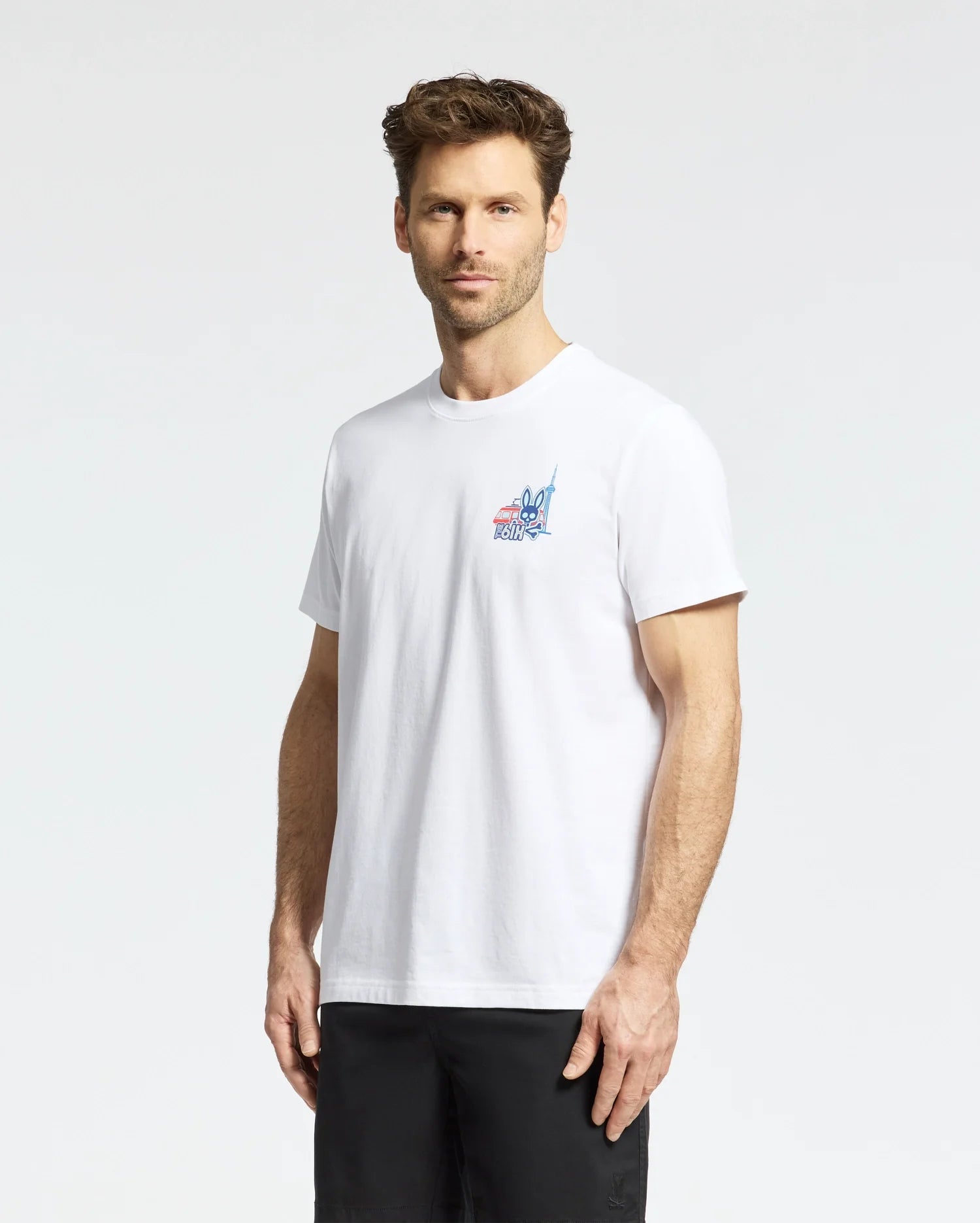 A man with short brown hair and a beard is wearing a Psycho Bunny MENS TORONTO TEE - B6U383F200, featuring a small colorful graphic on the left chest, along with black shorts, standing against a plain light background. (2)