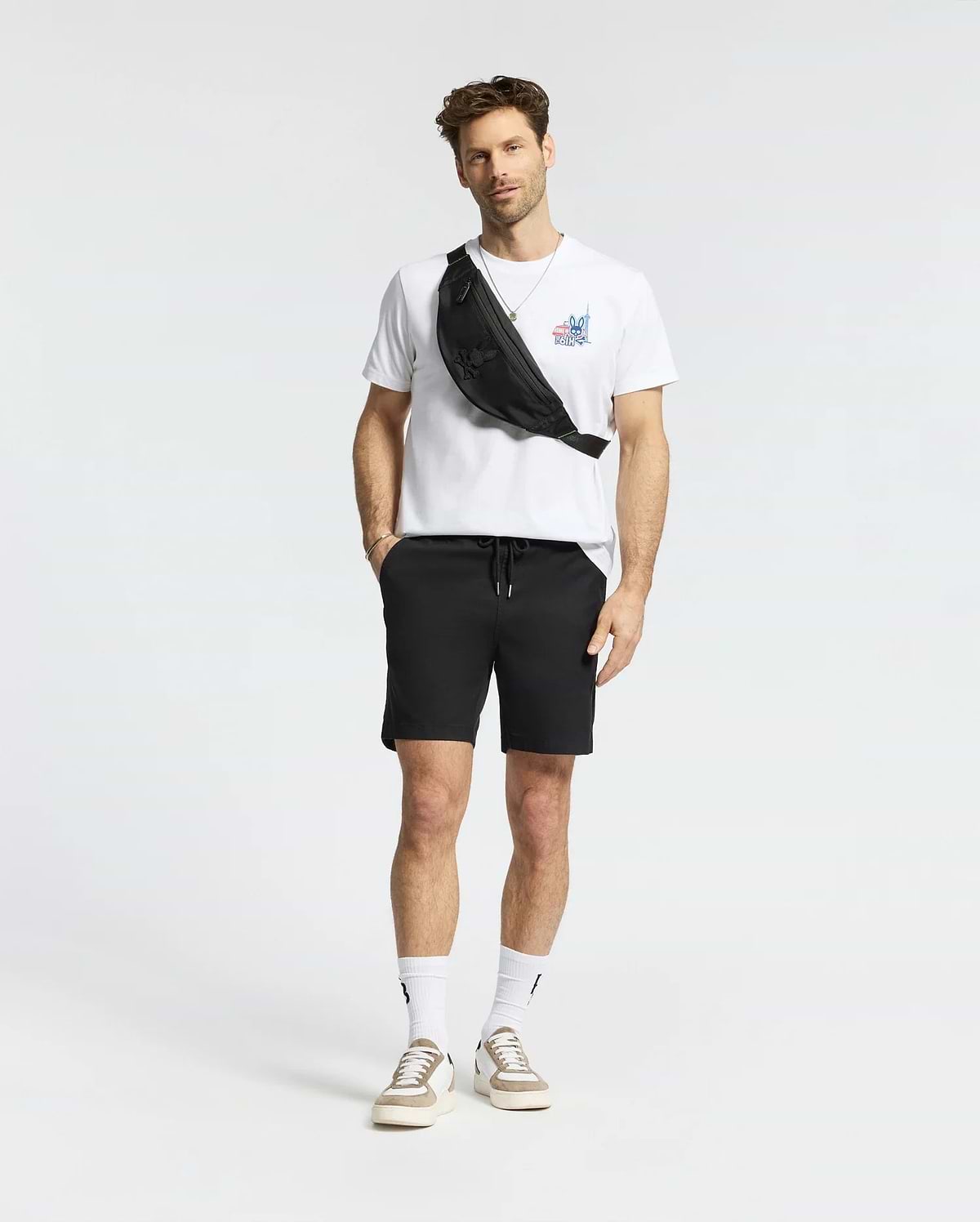 A man with short hair and a light beard stands against a white background, wearing the Psycho Bunny MENS TORONTO TEE - B6U383F200 in soft Pima cotton with bold graphics, black shorts, white socks, beige sneakers, and a black crossbody bag. (5)