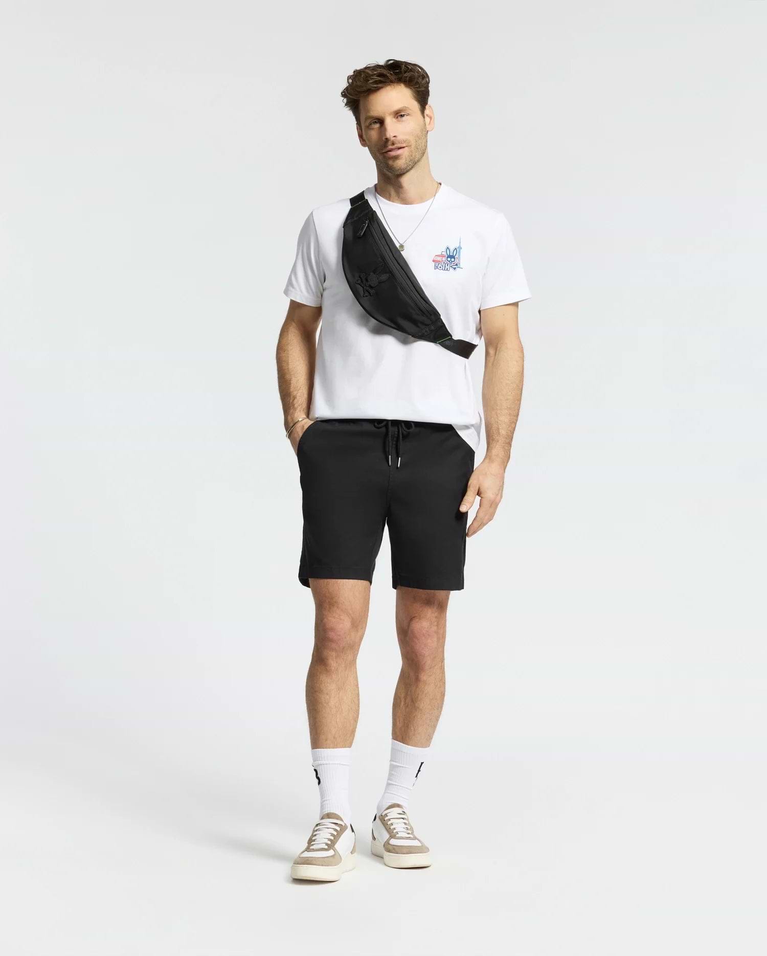 A man with short hair and a light beard stands against a white background, wearing the Psycho Bunny MENS TORONTO TEE - B6U383F200 in soft Pima cotton with bold graphics, black shorts, white socks, beige sneakers, and a black crossbody bag. (5)
