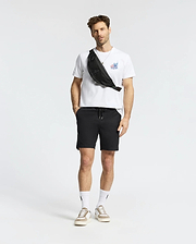 A man with short hair and a light beard stands against a white background, wearing the Psycho Bunny MENS TORONTO TEE - B6U383F200 in soft Pima cotton with bold graphics, black shorts, white socks, beige sneakers, and a black crossbody bag. (5)
