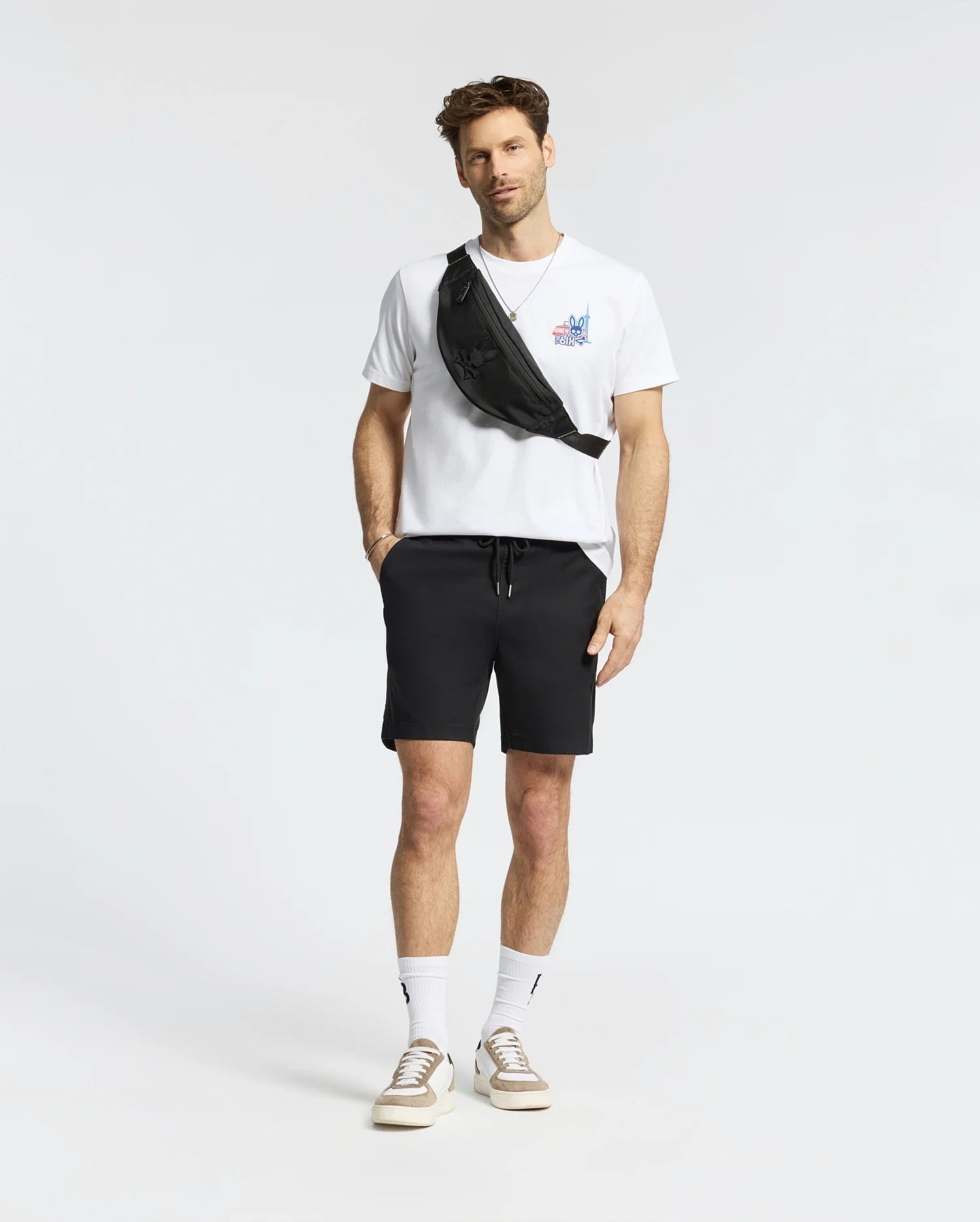 A man with short hair and a light beard stands against a white background, wearing the Psycho Bunny MENS TORONTO TEE - B6U383F200 in soft Pima cotton with bold graphics, black shorts, white socks, beige sneakers, and a black crossbody bag. (5)