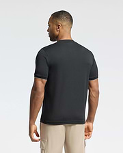 A man faces away wearing a Psycho Bunny MENS BRYCE PERFORMANCE TEE in black with white trim, made from pique fabric, paired with beige cargo shorts. The background is light gray.