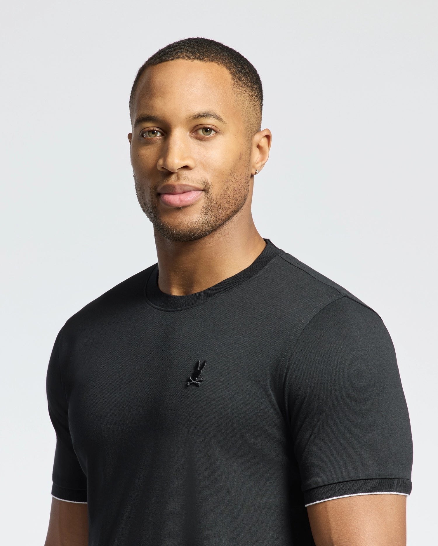 A man wearing the Psycho Bunny MENS BRYCE PERFORMANCE TEE - B6U490F200, made of Modal-Pima Cotton with a small logo, stands against a light gray background, looking at the camera with a neutral expression.