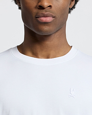 A close-up shows a person wearing the Psycho Bunny MENS BRYCE PERFORMANCE TEE - B6U490F200, a slim-fit, white T-shirt crafted from a luxurious Modal-Pima cotton blend, adorned with an embroidered rabbit and crossed bones logo. The plain white background accentuates the exquisite design.