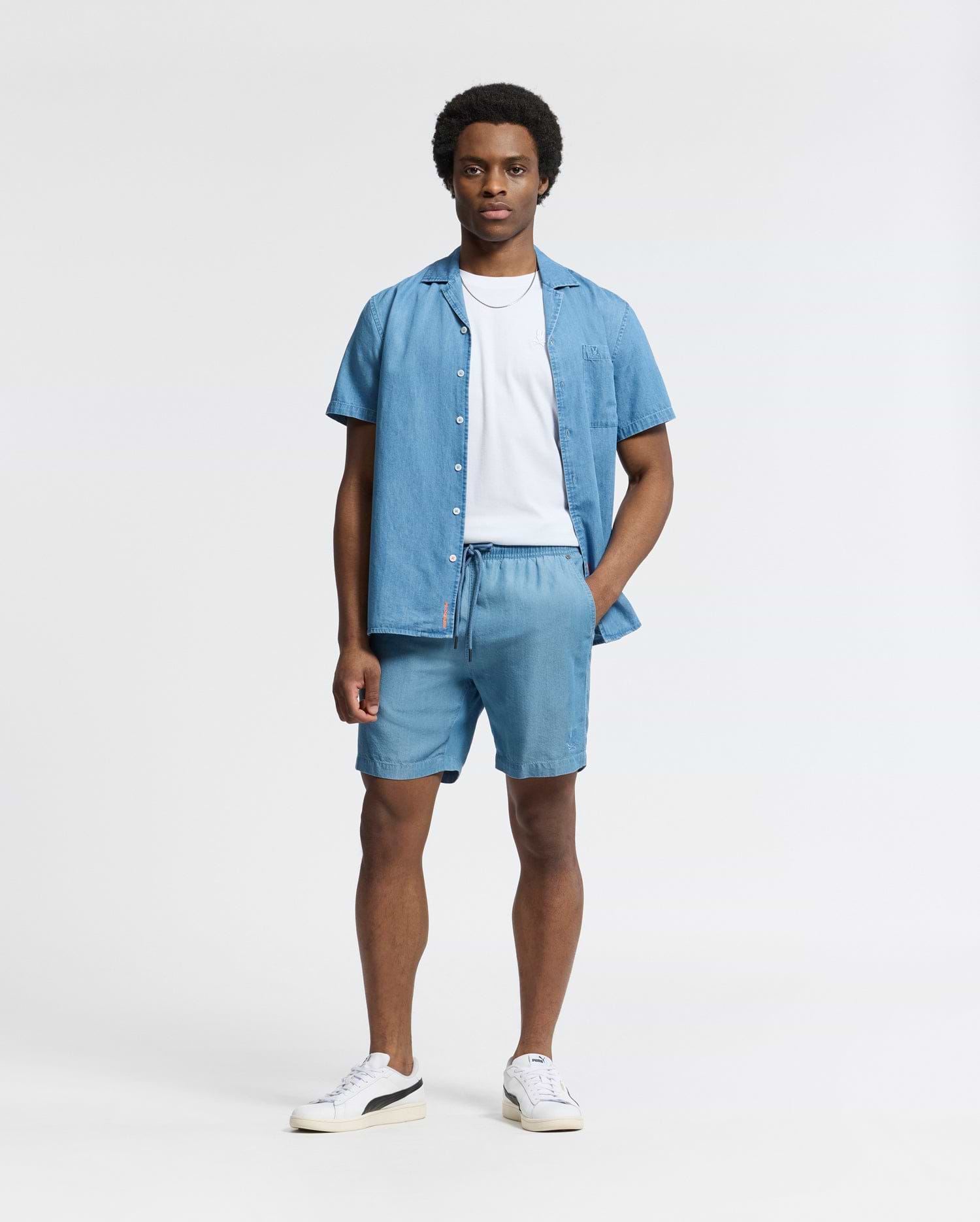 Against a light background, a person wears the Psycho Bunny MENS BRYCE PERFORMANCE TEE - B6U490F200, layered under a light blue slim-fit Modal-Pima cotton blend button-up shirt and matching shorts. They complete the look with white sneakers featuring black accents and stand with one hand in their pocket.
