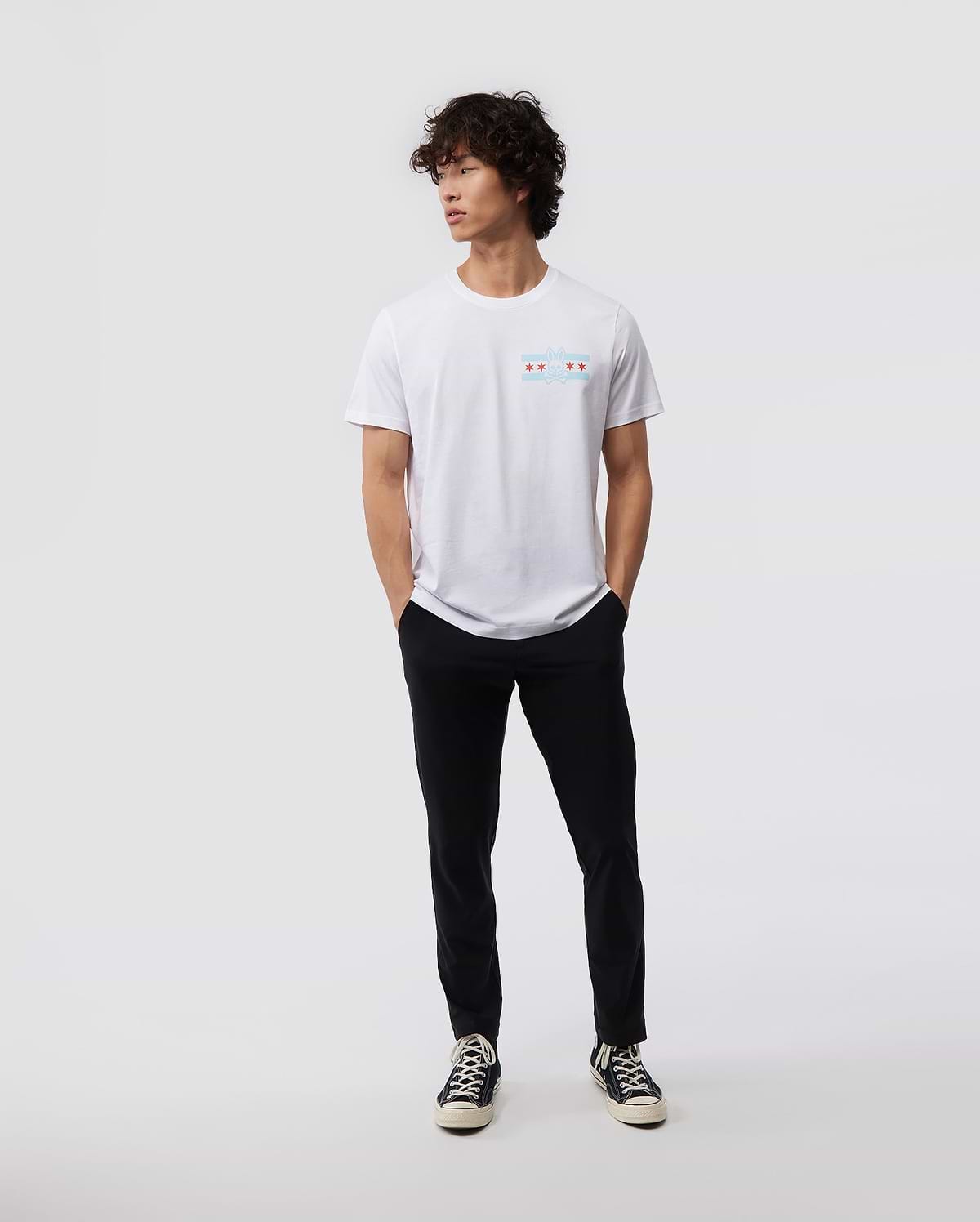 A person with curly hair stands casually against a plain background, wearing a Psycho Bunny MENS CHICAGO CITY TEE - B6U551W1PC, paired with black pants and classic black and white sneakers. Their hands rest in their pockets as they gaze to the side.