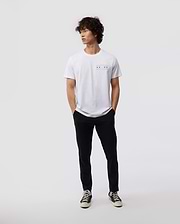 A person with curly hair stands casually against a plain background, wearing a Psycho Bunny MENS CHICAGO CITY TEE - B6U551W1PC, paired with black pants and classic black and white sneakers. Their hands rest in their pockets as they gaze to the side.