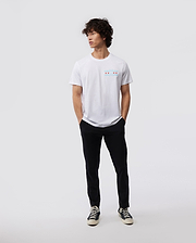 A person with curly hair stands casually against a plain background, wearing a Psycho Bunny MENS CHICAGO CITY TEE - B6U551W1PC, paired with black pants and classic black and white sneakers. Their hands rest in their pockets as they gaze to the side.
