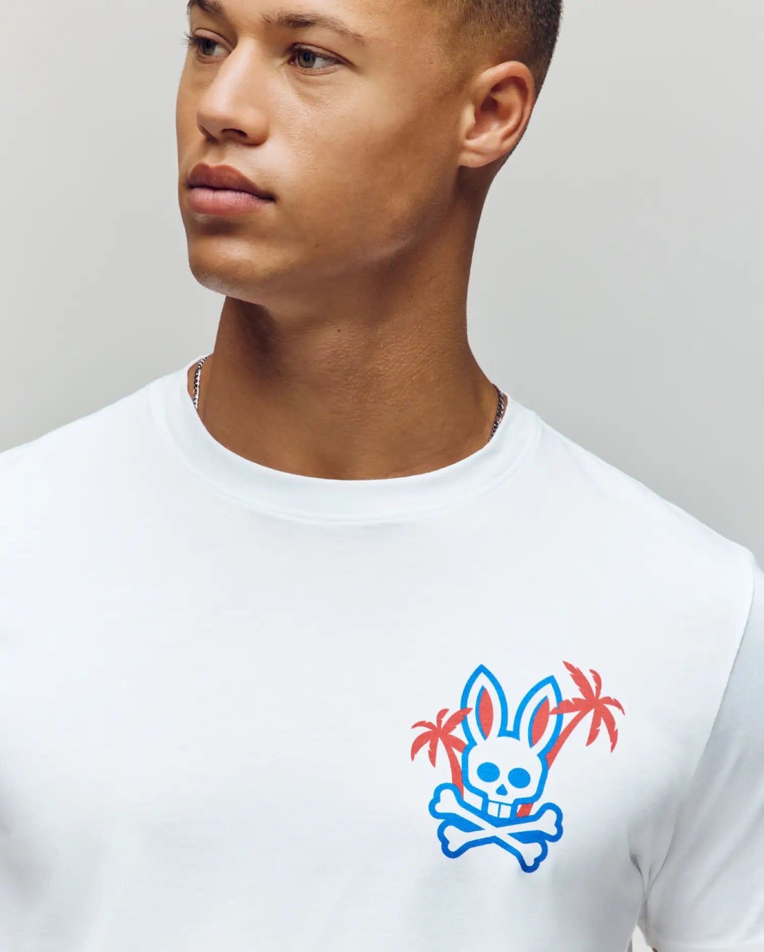 Wearing the Psycho Bunny MENS LOS ANGELES CITY TEE - B6U861A2PC, crafted from soft Pima cotton, a man features a retro Dodgers aesthetic with a skull and crossbones in blue and pink on neutral backdrop. (3)