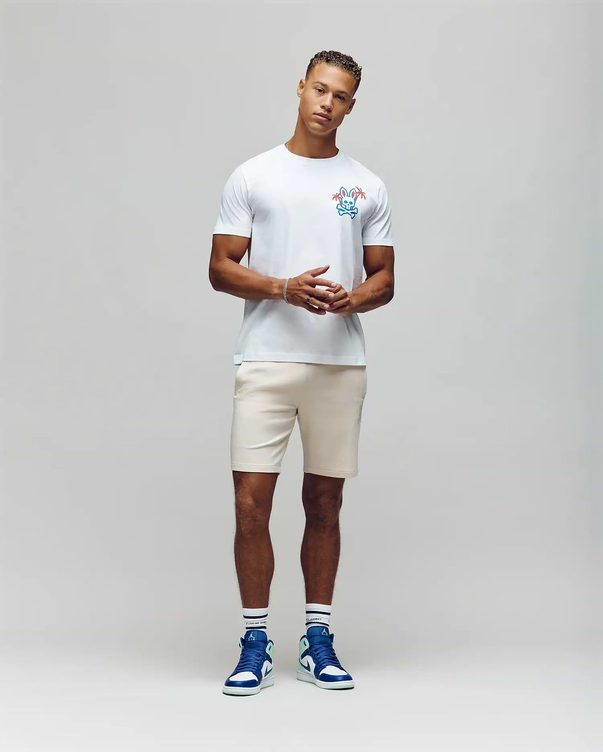 A man stands against a plain backdrop wearing the MENS LOS ANGELES CITY TEE by Psycho Bunny with a colorful chest graphic, light shorts, and blue high-top sneakers. His hands are clasped in front of him, effortlessly channeling a retro Dodgers aesthetic. (4)