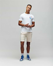 A man stands against a plain backdrop wearing the MENS LOS ANGELES CITY TEE by Psycho Bunny with a colorful chest graphic, light shorts, and blue high-top sneakers. His hands are clasped in front of him, effortlessly channeling a retro Dodgers aesthetic. (4)