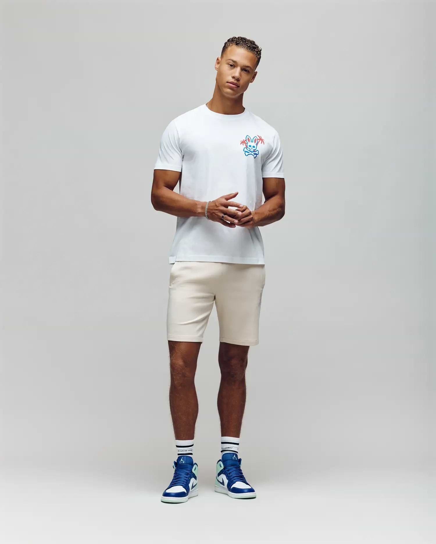 A man stands against a plain backdrop wearing the MENS LOS ANGELES CITY TEE by Psycho Bunny with a colorful chest graphic, light shorts, and blue high-top sneakers. His hands are clasped in front of him, effortlessly channeling a retro Dodgers aesthetic. (4)