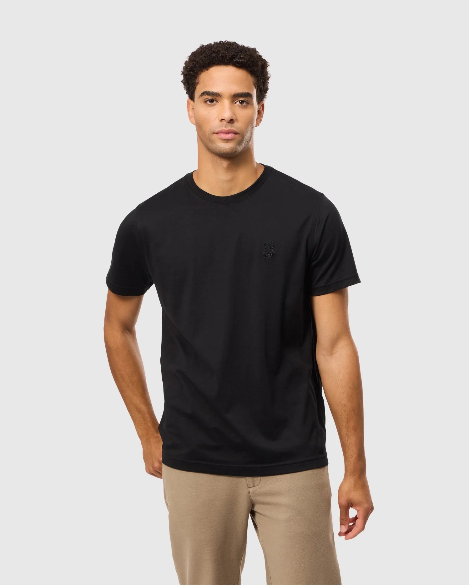 A man with short curly hair is standing against a plain light gray background, wearing the Psycho Bunny MENS JAMES PREMIUM CREW NECK TEE - B6U952D200, made of Pima cotton, and light brown pants. His left hand is in his pocket, and he has a neutral expression on his face.