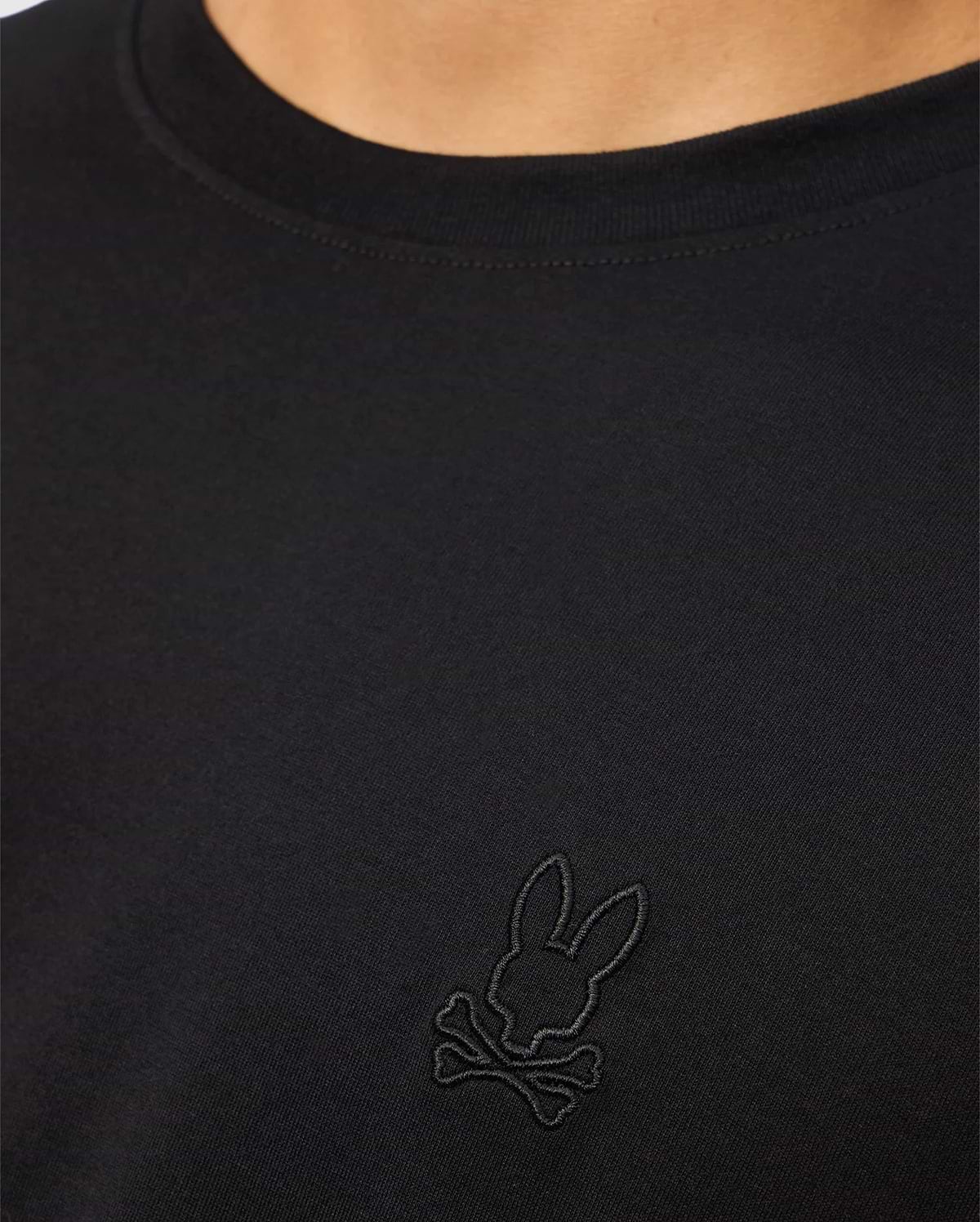 A close-up of a person showcasing the MENS JAMES PREMIUM CREW NECK TEE - B6U952D200 by Psycho Bunny. This black Pima cotton shirt features an embroidered black logo on the chest, depicting a bunny head with crossbones underneath. The person's neck and part of the shoulder are visible.