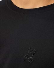 A close-up of a person showcasing the MENS JAMES PREMIUM CREW NECK TEE - B6U952D200 by Psycho Bunny. This black Pima cotton shirt features an embroidered black logo on the chest, depicting a bunny head with crossbones underneath. The person's neck and part of the shoulder are visible.
