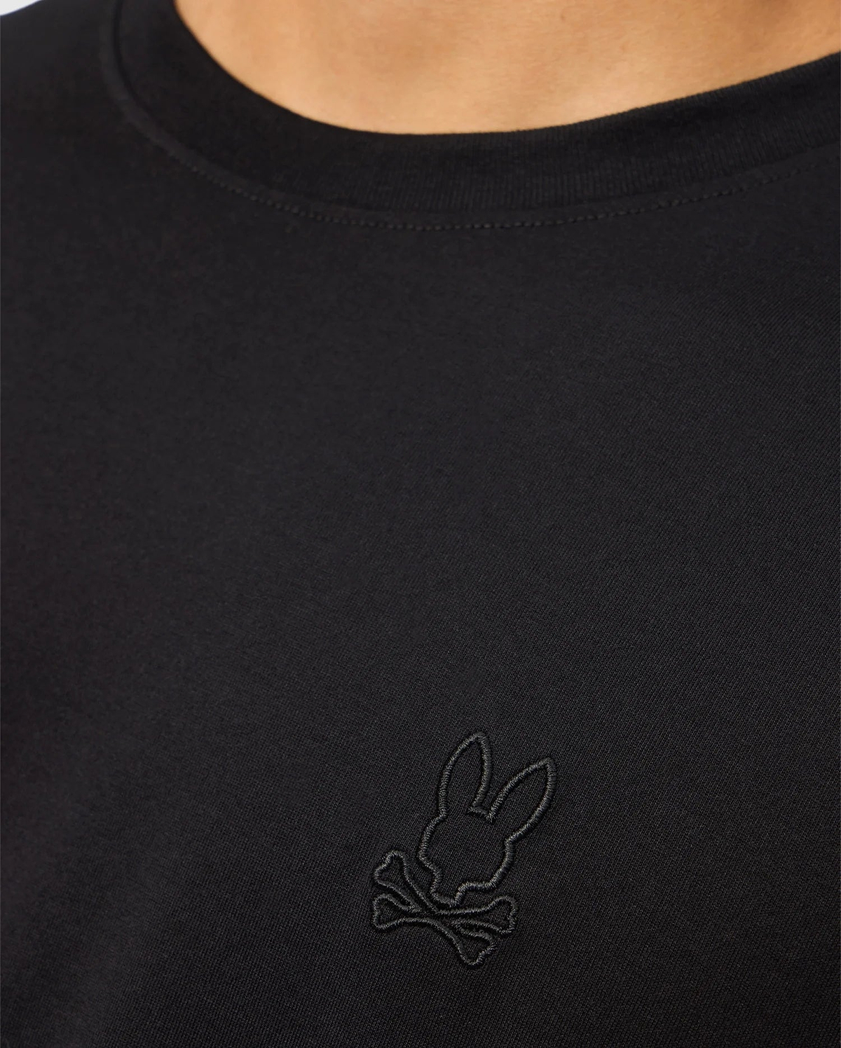 A close-up of a person showcasing the MENS JAMES PREMIUM CREW NECK TEE - B6U952D200 by Psycho Bunny. This black Pima cotton shirt features an embroidered black logo on the chest, depicting a bunny head with crossbones underneath. The person's neck and part of the shoulder are visible.