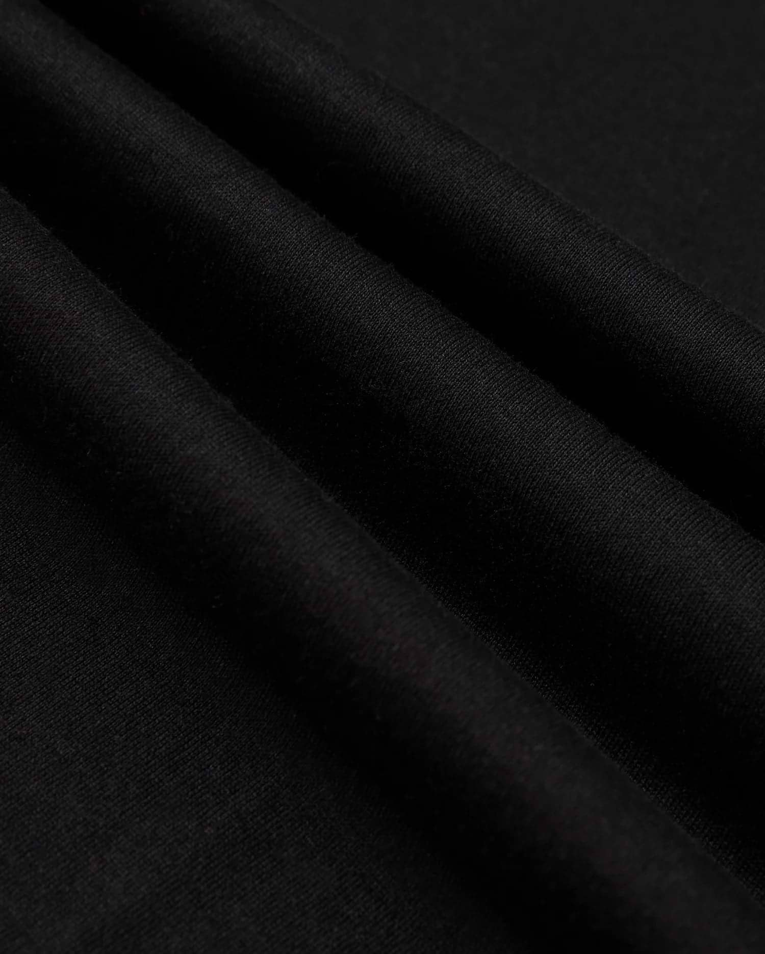 Close-up of the neatly folded black Pima cotton fabric with a soft texture. Three prominent folds create shadows, emphasizing the smooth material. The image highlights the quality and richness of Psycho Bunny's MENS JAMES PREMIUM CREW NECK TEE - B6U952D200, showcasing its sleek and elegant appearance.