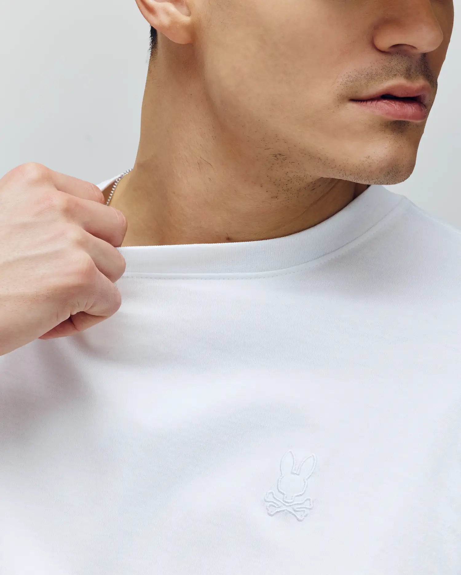 A person wearing the Psycho Bunny MENS JAMES PREMIUM CREW NECK TEE (B6U952D200), made from Premium Pima cotton with an embroidered rabbit and crossbones logo, holds the collar near their neck. The lower half of their face is visible.