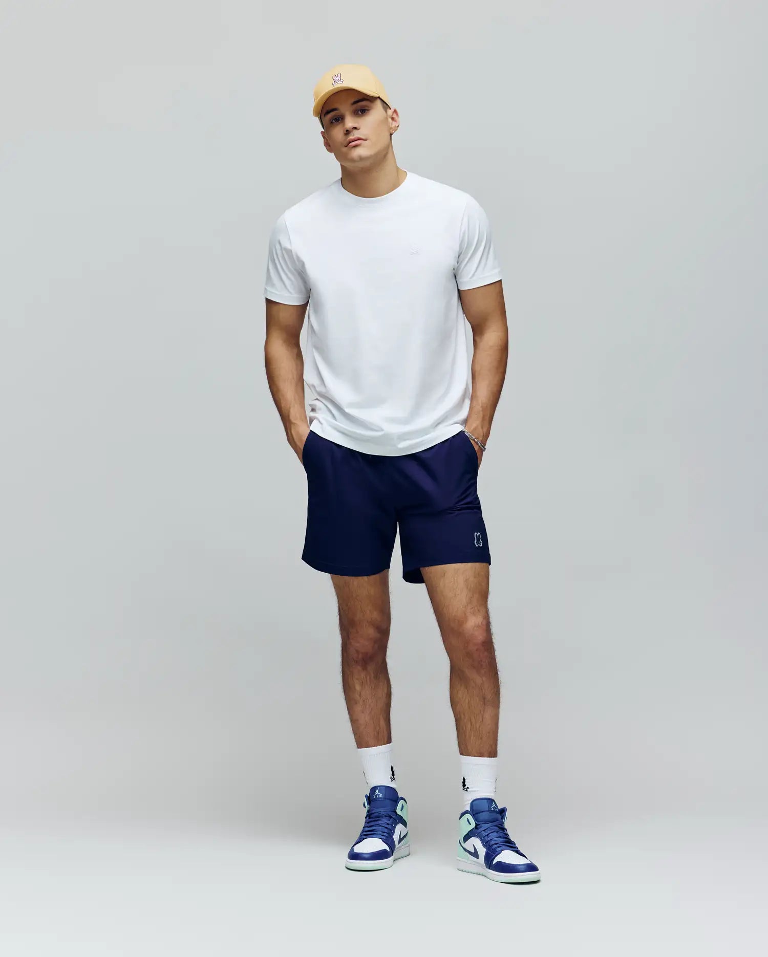 A man stands against a light gray background wearing a yellow cap, navy shorts, white crew socks, blue and white sneakers, and the Psycho Bunny MENS JAMES PREMIUM CREW NECK TEE (B6U952D200), with hands in his pockets.