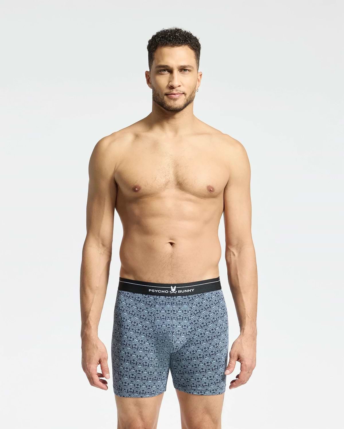 A man with short curly hair stands against a plain white background, wearing Psycho Bunny's Mens 2 Pack Boxer Briefs (B6V481F200) featuring a black waistband. He looks directly at the camera, showcasing the comfort and style of the breathable stretch Pima cotton.