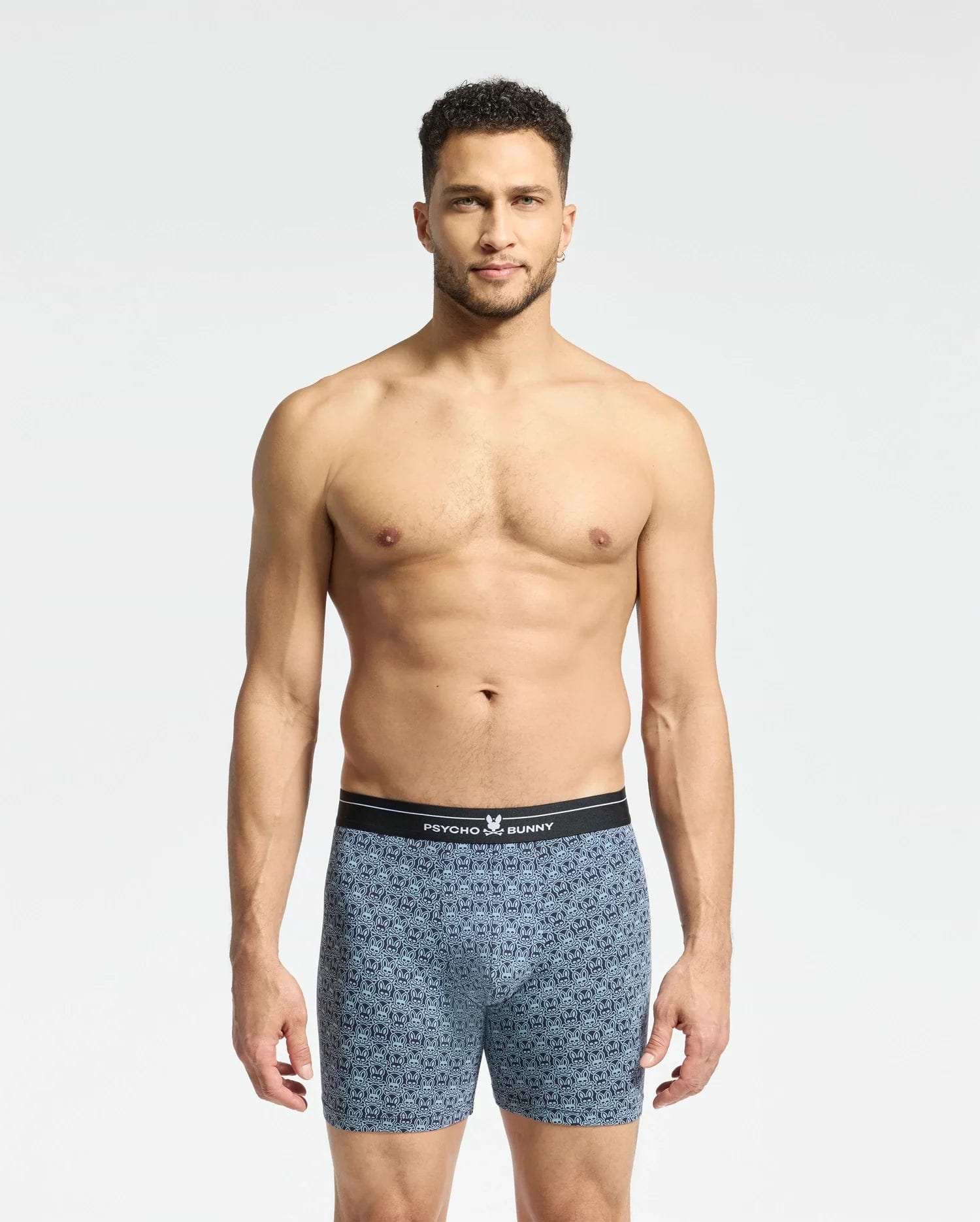 A man with short curly hair stands against a plain white background, wearing Psycho Bunny's Mens 2 Pack Boxer Briefs (B6V481F200) featuring a black waistband. He looks directly at the camera, showcasing the comfort and style of the breathable stretch Pima cotton.