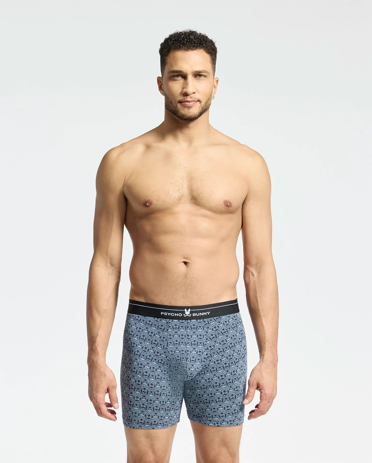 A man with short curly hair stands against a plain white background, wearing Psycho Bunny's Mens 2 Pack Boxer Briefs (B6V481F200) featuring a black waistband. He looks directly at the camera, showcasing the comfort and style of the breathable stretch Pima cotton.