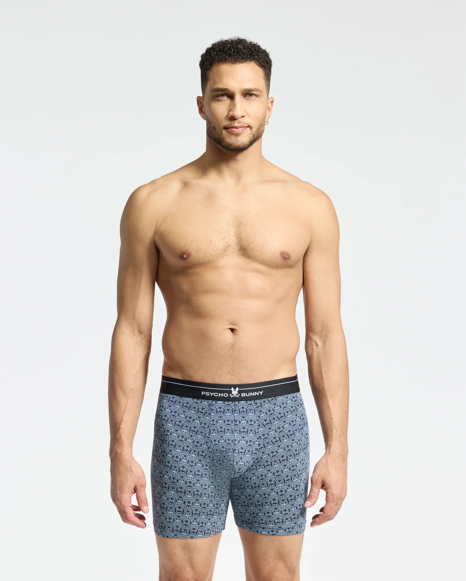 A man with short curly hair stands against a plain white background, wearing Psycho Bunny's Mens 2 Pack Boxer Briefs (B6V481F200) featuring a black waistband. He looks directly at the camera, showcasing the comfort and style of the breathable stretch Pima cotton.