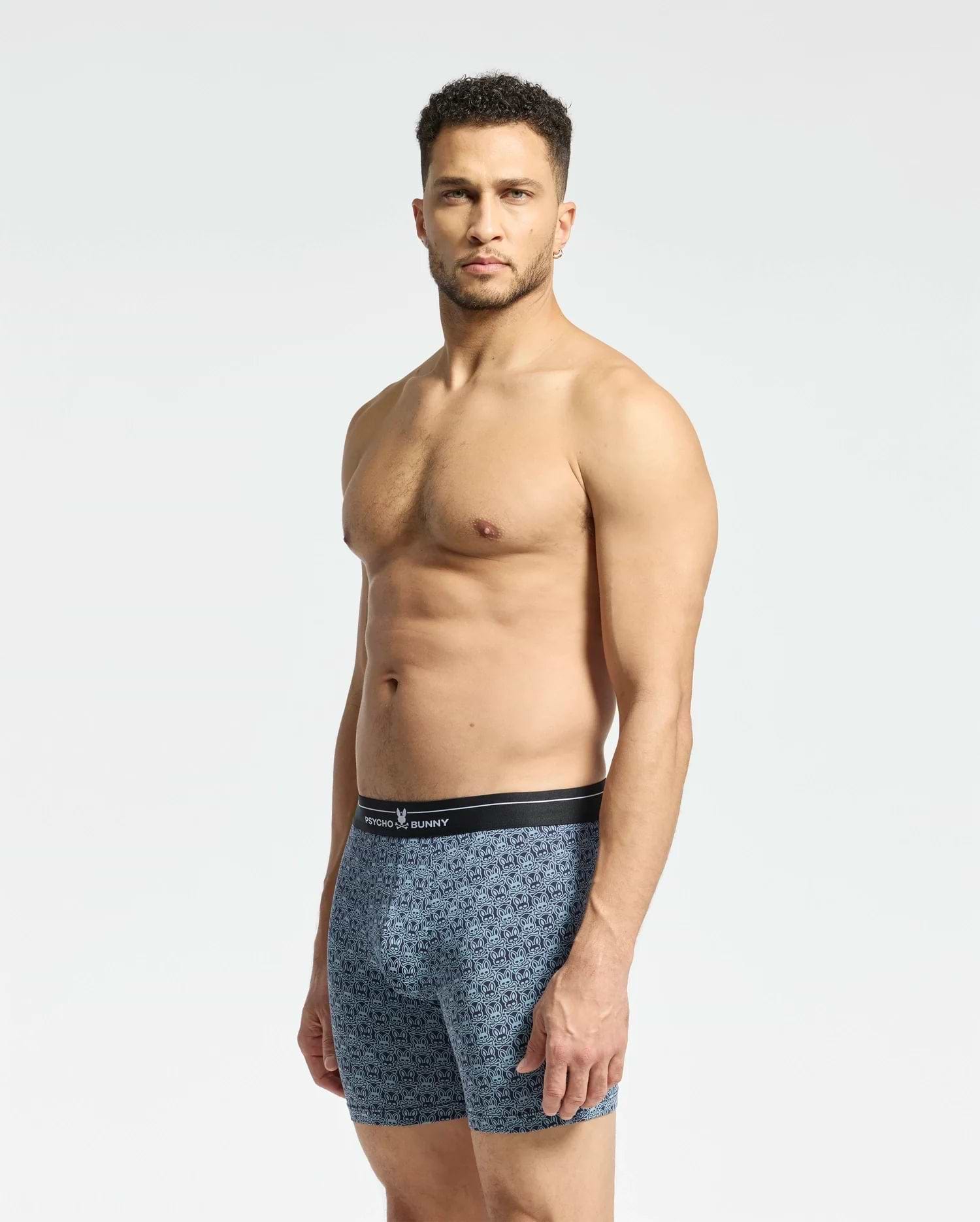 A shirtless man stands against a plain background, wearing Psycho Bunny MENS 2 PACK BOXER BRIEF - B6V481F200, featuring blue patterns and a black waistband made from breathable fabric. He looks directly at the camera.