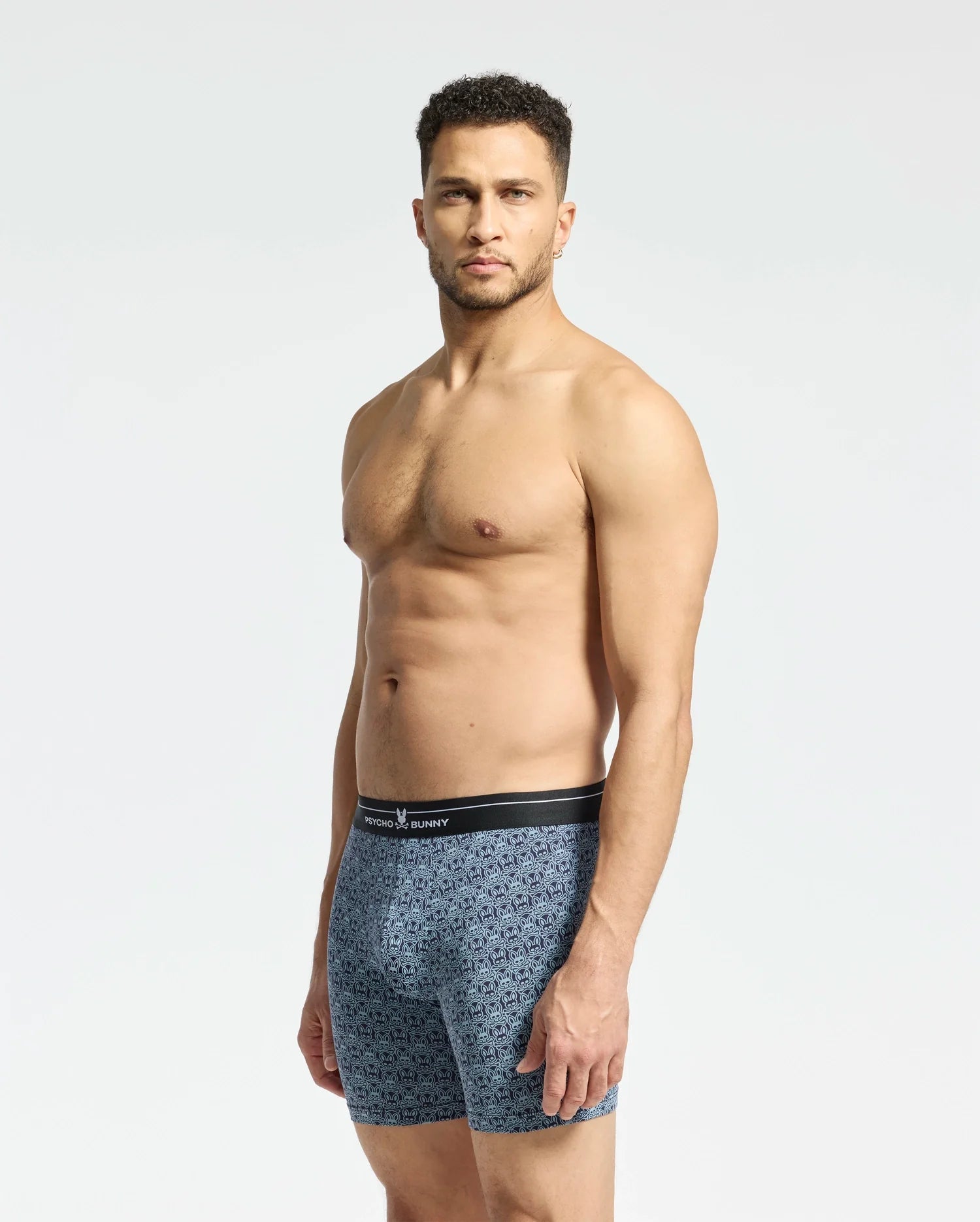 A shirtless man stands against a plain background, wearing Psycho Bunny MENS 2 PACK BOXER BRIEF - B6V481F200, featuring blue patterns and a black waistband made from breathable fabric. He looks directly at the camera.